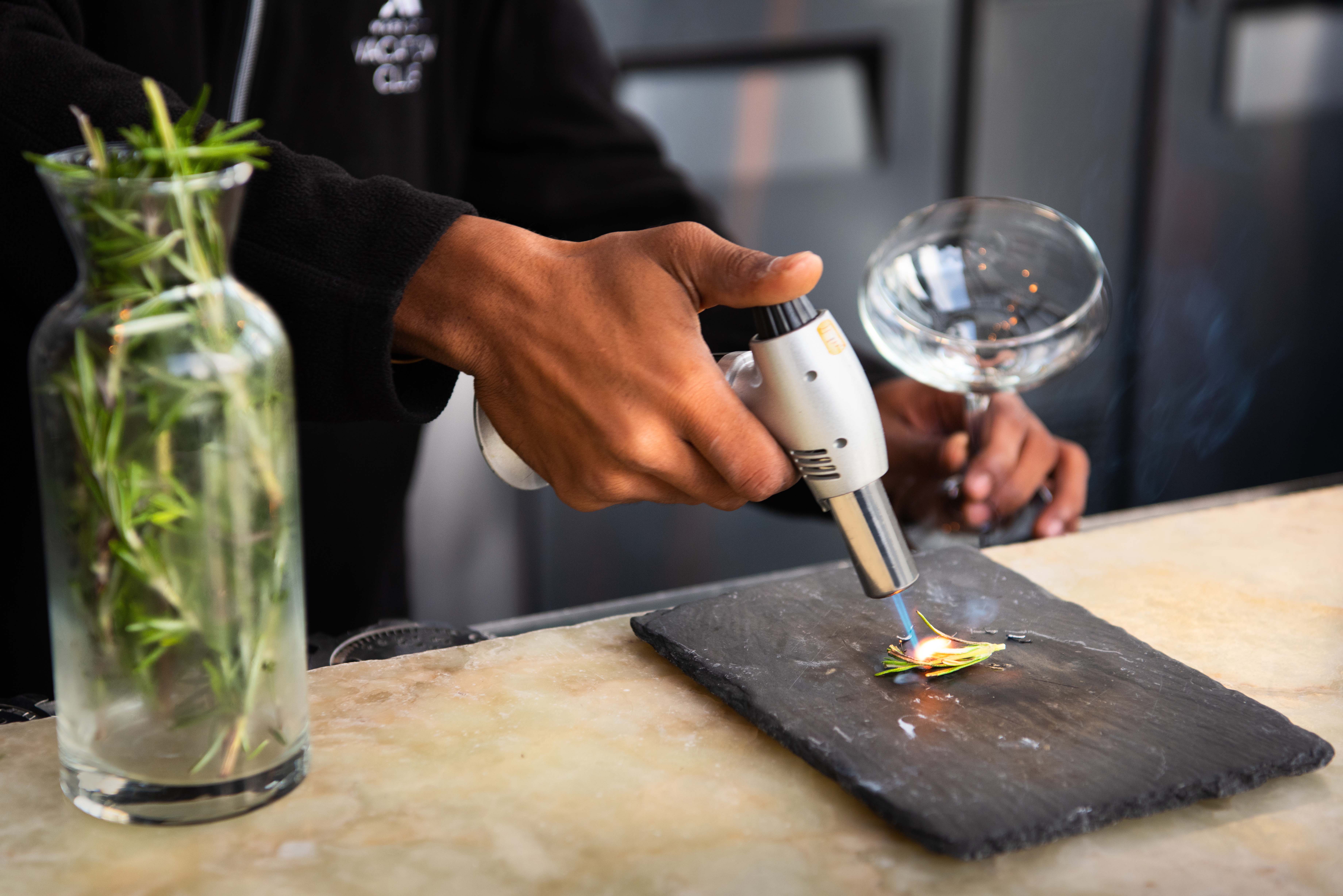 Man making cocktail garnish using a hand torch on green herbs